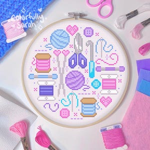 以下が含まれることがあります： ピンクと白の背景に、ピンクのハートで囲まれた、糸巻き、ハサミ、針、糸巻きなど、さまざまな縫製と編みの用品がカラフルなクロスステッチでデザインされた白い刺繍枠。