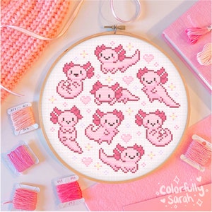May include: A pink and white cross-stitch embroidery hoop with a design of eight pink axolotls with hearts and stars. The hoop is on a pink surface with pink yarn and embroidery floss.