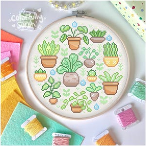 以下が含まれることがあります： 様々な緑色のトーンの鉢植えの植物のデザインが施されたクロスステッチ刺繍フープ。植物は小さな青い雨滴で囲まれています。フープは、カラフルな布切れと刺繍糸のある白い表面の上にあります。