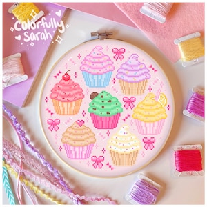 May include: A pink embroidery hoop with a cross-stitch design of colorful cupcakes. The cupcakes are decorated with sprinkles, cherries, chocolate chips, and lemon slices. The hoop is surrounded by colorful embroidery floss.