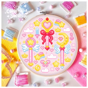 Puede incluir: Un patrón de punto de cruz rosa y amarillo con un diseño caprichoso de estrellas, corazones, coronas y varitas. El patrón está enmarcado en un aro de madera.