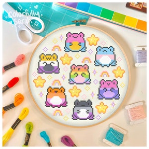 Peut inclure: Un cercle de broderie circulaire avec un fond blanc et des grenouilles pixelisées colorées. Les grenouilles sont disposées en cercle autour du cercle et sont entourées d'étoiles et d'arcs-en-ciel. Le cercle est en bois et a une finition naturelle.
