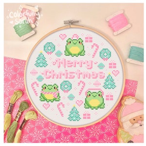 Puede incluir: Un aro de punto de cruz blanco con un diseño navideño rosa y verde con tres ranas, dos con gorros de Papá Noel, y el texto "Feliz Navidad".