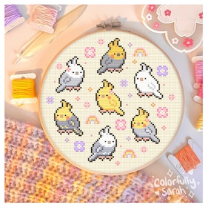 May include: A cross-stitch embroidery hoop featuring a design of eight yellow and grey cockatiels with pink flowers and rainbows on a cream background. The hoop is on a knitted fabric with a pink, yellow, and grey colour scheme.