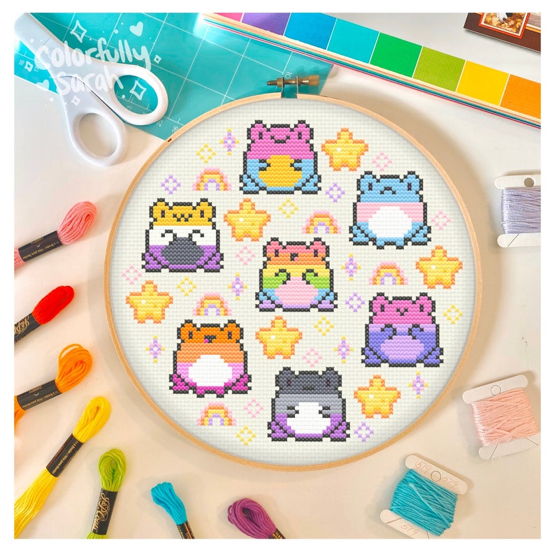 May include: A circular embroidery hoop with a white background and colourful pixelated frogs. The frogs are arranged in a circle around the hoop and are surrounded by stars and rainbows. The hoop is made of wood and has a natural finish.