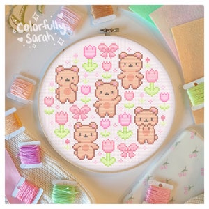 May include: A white embroidery hoop with a pink and green cross-stitch design featuring six brown teddy bears, pink tulips, and green leaves. The design is surrounded by a white border.
