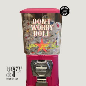 May include: Pink gumball machine with a clear plastic container filled with colorful gumballs. The machine has a sticker that says "Don't Worry Doll you got it" with a red star. The machine has a silver coin slot and a lever to dispense the gumballs.