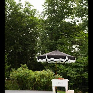 May include: A black and white fringed patio umbrella stands over a white rolling cart next to a pool. The cart has a beverage cooler and a tote bag. Lush green trees fill the background.