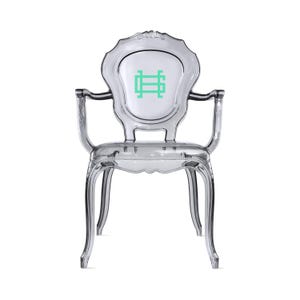 May include: A clear, transparent armchair with a classic design. The chair features armrests and a backrest with a white center panel. A green logo is visible on the backrest. The chair is made of a clear, durable material.
