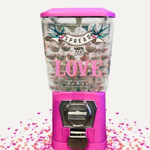 May include: A bright pink gumball machine with a clear top filled with gumballs. The machine has the words "SPREAD The LOVE BOMBS" printed on the front, with a banner and swallows graphic.