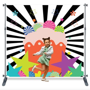 May include: A colorful backdrop with a large cupcake design, black and white stripes, and colorful stars. A young girl is standing in front of the backdrop, wearing a green dress and a white jacket.