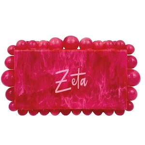 May include: A bright pink clutch bag with a marbled effect and a white border. The bag has a rectangular shape and is decorated with a row of round beads around the perimeter. The word "Zeta" is written in white letters on the front of the bag.