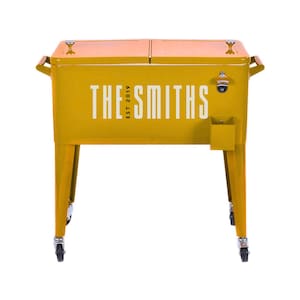May include: A yellow metal cooler with the text "THE SMITHS EST. 2019" in white. The cooler has a bottle opener, a small storage compartment, and wooden handles. It stands on four legs with wheels.