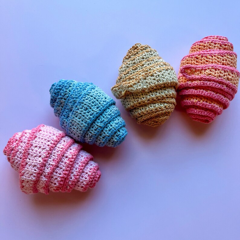 May include: Four crocheted egg-shaped ornaments in different colors: pink, blue, yellow, and orange. The ornaments are made with a spiral pattern and are arranged in a row on a light purple background.