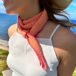 May include: A pink and orange crocheted scarf with a knotted design. The scarf is worn around the neck of a person wearing a white tank top.