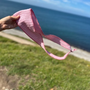 May include: A pink crocheted scarf is held in a hand against a backdrop of a blue ocean and a green grassy hill.