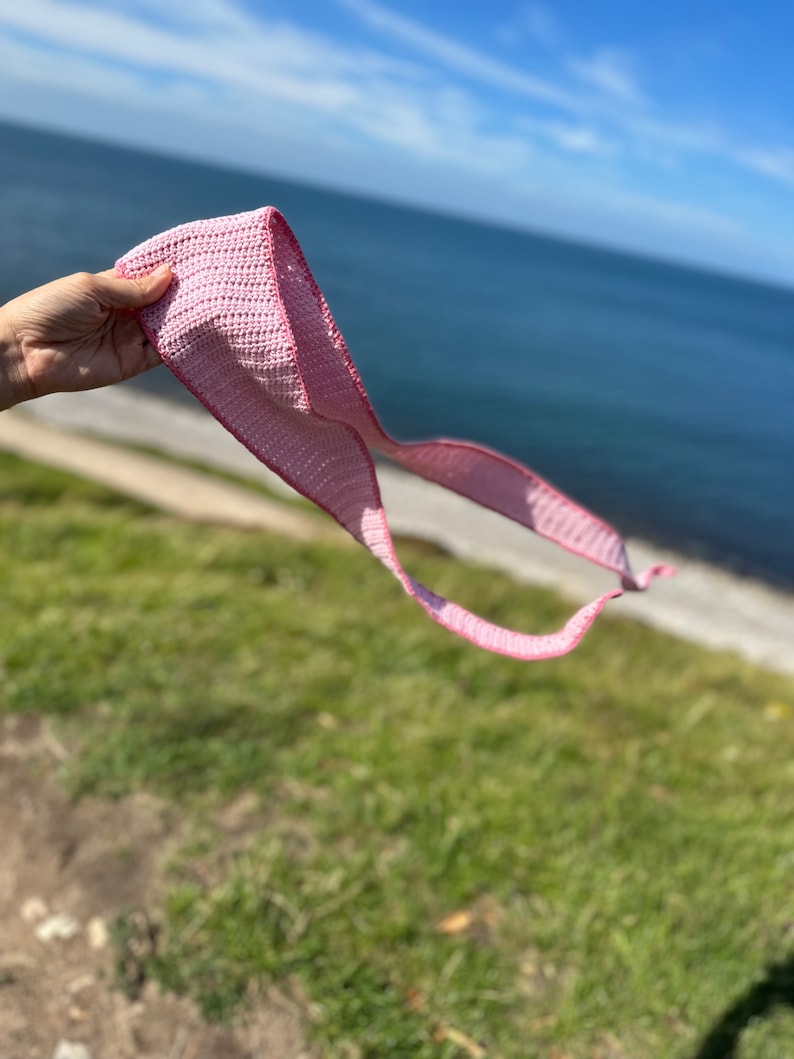 May include: A pink crocheted scarf is held in a hand against a backdrop of a blue ocean and a green grassy hill.