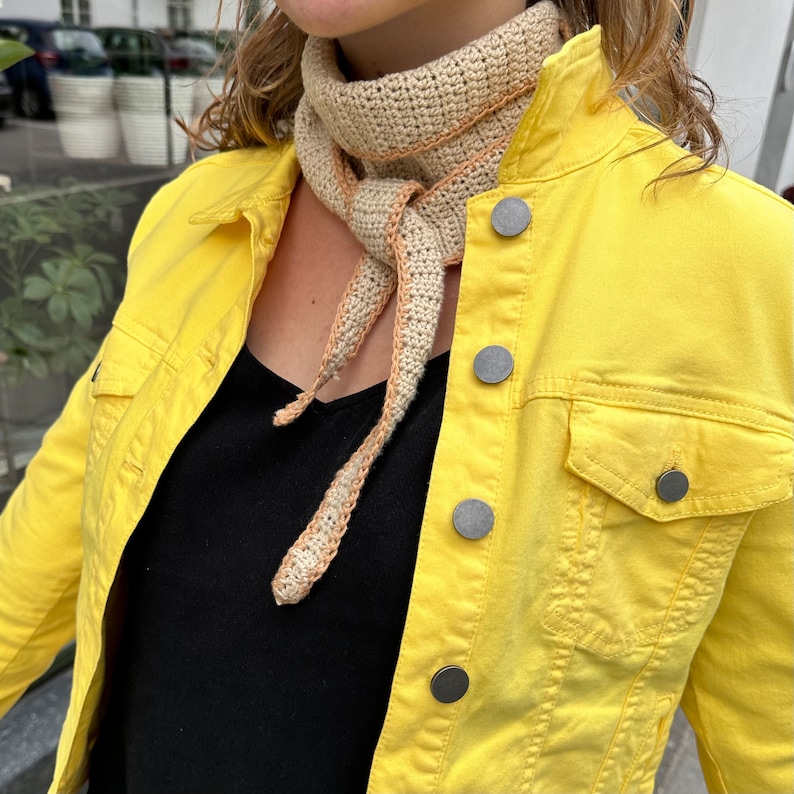 May include: A yellow denim jacket with a crocheted scarf in beige and brown. The scarf is tied in a knot at the neck and hangs down the front of the jacket.