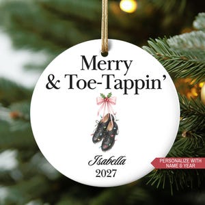 May include: White round Christmas ornament with the text "Merry & Toe-Tappin'" and a pair of black tap shoes. The ornament has a gold string for hanging and the name "Isabella" and the year "2027" are printed on the ornament.