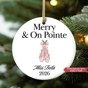 May include: White round Christmas ornament with the text "Merry & On Pointe" and a watercolor illustration of pink ballet shoes. The ornament also includes the name "Miss Bella" and the year "2026".
