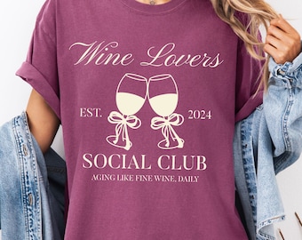 May include: A maroon t-shirt with the text "Wine Lovers Social Club" and two wine glasses. The shirt includes the text "EST. 2024" and "Aging Like Fine Wine, Daily". The shirt is a comfortable fit.