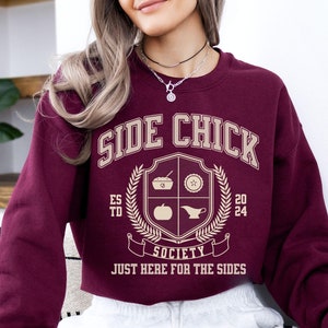 May include: Burgundy cropped sweatshirt with the text "Side Chick Society" and a crest featuring a pumpkin, pie, gravy boat, and bowl. The text below the crest reads "Just Here For The Sides."