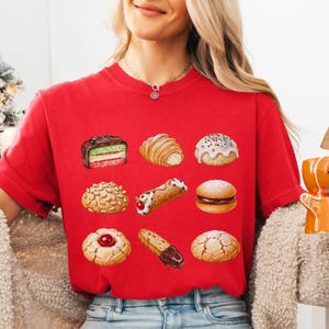 May include: A red t-shirt featuring a variety of illustrated pastries, including a croissant, a cannoli, and a cookie with a red center. The shirt is paired with blue jeans. A gingerbread man mug is held in the hand.