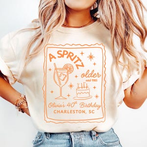 May include: Cream-colored t-shirt with an orange graphic. The graphic includes the text "A Spritz older estd 1985" with a cocktail illustration, cake, and the text "Olivia's 40th Birthday Charleston, SC".