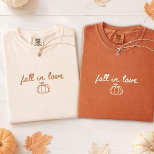 May include: Two short-sleeve t-shirts, one cream and one burnt orange, each featuring the text fall in love and a simple pumpkin line drawing. Both shirts have a crew neckline and a Comfort Colors brand label inside the collar area.