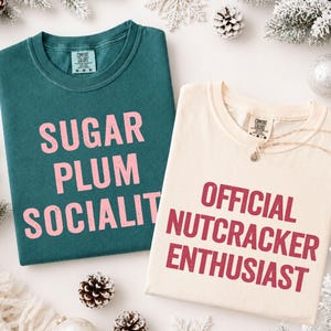 May include: A dark teal t-shirt with the pink text "SUGAR PLUM SOCIALITE" and a cream-colored t-shirt with the dark red text "OFFICIAL NUTCRACKER ENTHUSIAST". Both shirts feature a crew neckline and a Comfort Colors brand label at the inner collar.