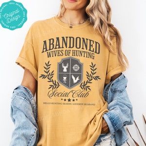 May include: A mustard yellow t-shirt with the text "Abandoned Wives of Hunting Social Club" in black. The shirt has a gray shield with a deer, target, wine glass, and bird inside. The text "Hello Hunting Season. Goodbye Husband" is below the shield.
