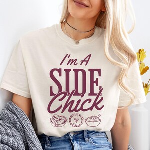 May include: A white t-shirt with the text "I'm a Side Chick" in maroon lettering. The text is accompanied by three maroon illustrations of Thanksgiving side dishes: a bowl of green beans, a pie, and a bowl of mashed potatoes.