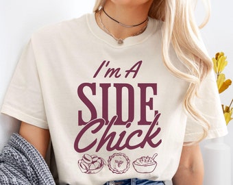 May include: A white t-shirt with the text "I'm a Side Chick" in maroon lettering. The text is accompanied by three maroon illustrations of Thanksgiving side dishes: a bowl of green beans, a pie, and a bowl of mashed potatoes.