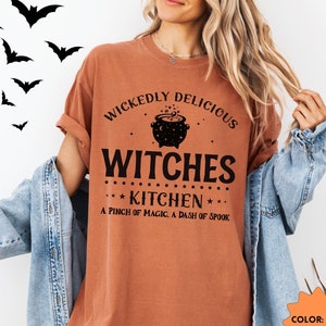 May include: A rust-colored t-shirt with a black graphic that says "Wickedly Delicious Witches Kitchen A Pinch of Magic. A Dash of Spook".