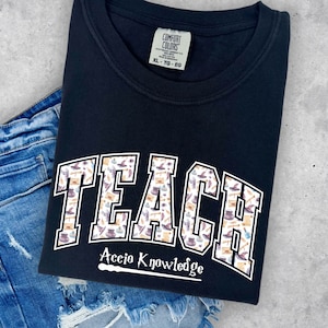 May include: Black t-shirt with a white "TEACH" graphic featuring a pattern of wizarding objects. The text "Accio Knowledge" is below the graphic with a white wand.