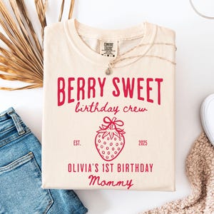 May include: Cream-colored t-shirt with the text "BERRY SWEET birthday crew" in red. A red strawberry graphic is below the text, with the words "EST. 2025" and "OLIVIA'S 1ST BIRTHDAY Mommy" also in red. The shirt is laid on a surface with jeans and a shoe.