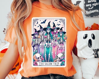 May include: A woman wearing an orange t-shirt with a graphic of a tarot card featuring a group of skeleton witches in colorful robes and hats. The card reads "The Salem Trip".