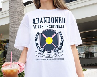 May include: White t-shirt with a graphic design that says "Abandoned Wives of Softball Social Club" with a softball and crossed bats in a crest. The text below the crest says "Hello Softball Season: Goodbye Husband."