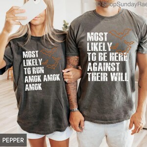 May include: Two matching dark gray t-shirts with white text and orange bat graphics. One shirt reads "MOST LIKELY TO RUN AMOK AMOK AMOK" and the other says "MOST LIKELY TO BE HERE AGAINST THEIR WILL".