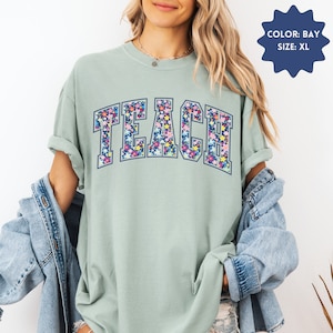 May include: A light green t-shirt with a floral print of the word "TEACH" in navy blue. The shirt is a relaxed fit and has a crew neck.