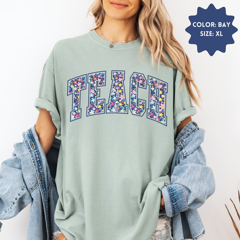 May include: A light green t-shirt with a floral print of the word "TEACH" in navy blue. The shirt is a relaxed fit and has a crew neck.