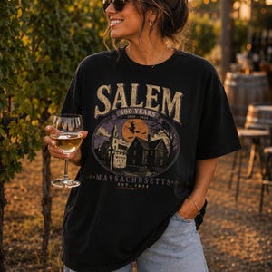May include: A black short-sleeve t-shirt featuring a graphic print with the text SALEM 400 YEARS 1626-2026, WITCH CITY 1693, MASSACHUSETTS, and EST. 1626. The design depicts a witch on a broomstick, a full moon, and historic buildings.