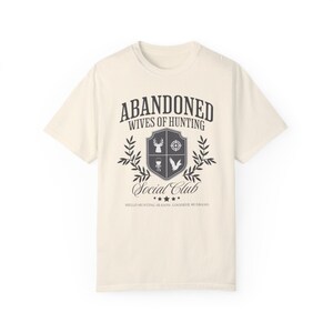May include: A beige t-shirt with a black graphic design that says "Abandoned Wives of Hunting Social Club" with a crest featuring a deer, a target, a bird, and a grill. The text below the crest says "Hello Hunting Season, Goodbye Husband."