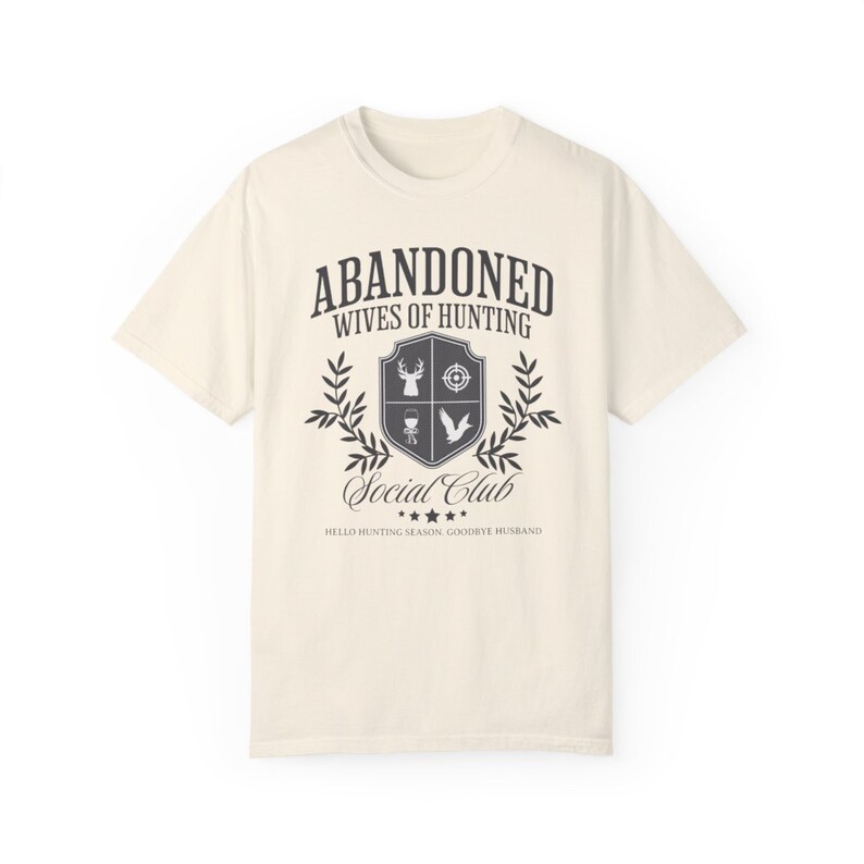 May include: A beige t-shirt with a black graphic design that says "Abandoned Wives of Hunting Social Club" with a crest featuring a deer, a target, a bird, and a grill. The text below the crest says "Hello Hunting Season, Goodbye Husband."
