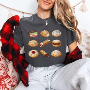 May include: Dark gray t-shirt featuring a watercolor illustration of various pastries, including a layered cake, croissant, and cannoli. The shirt has a crew neck and short sleeves. The model is wearing a red and black plaid shirt over the t-shirt.