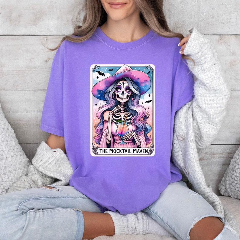 May include: Purple t-shirt with a graphic of a skeleton witch wearing a witch hat and holding a cocktail. The graphic is in a card-like frame with the text "The Mocktail Maven".