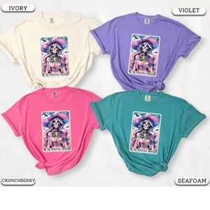 May include: Four different colored t-shirts featuring a colorful illustration of a skeleton wearing a witch's hat and holding a glass of liquid. The text "The Mocktail Maven" is printed below the illustration.