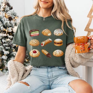 May include: An olive green t-shirt featuring watercolor illustrations of various pastries, including a croissant, a cannoli, and a layered cake. The person is holding a gingerbread man mug. The shirt is paired with light blue jeans and a cream-colored cardigan.