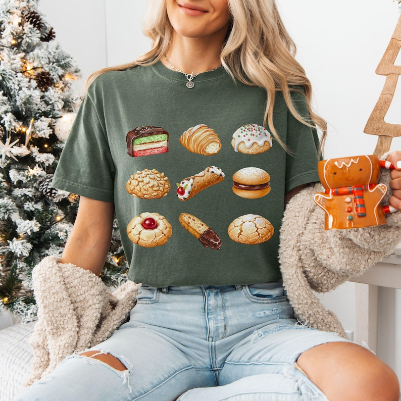 May include: An olive green t-shirt featuring watercolor illustrations of various pastries, including a croissant, a cannoli, and a layered cake. The person is holding a gingerbread man mug. The shirt is paired with light blue jeans and a cream-colored cardigan.