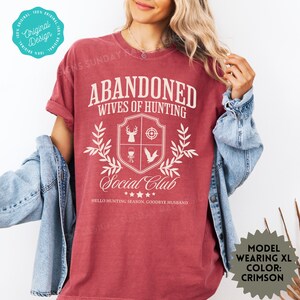 May include: A red t-shirt with the text "Abandoned Wives of Hunting Social Club" and a crest featuring a deer, a target, a wine glass, and a bird. The text "Hello Hunting Season, Goodbye Husband" is below the crest.